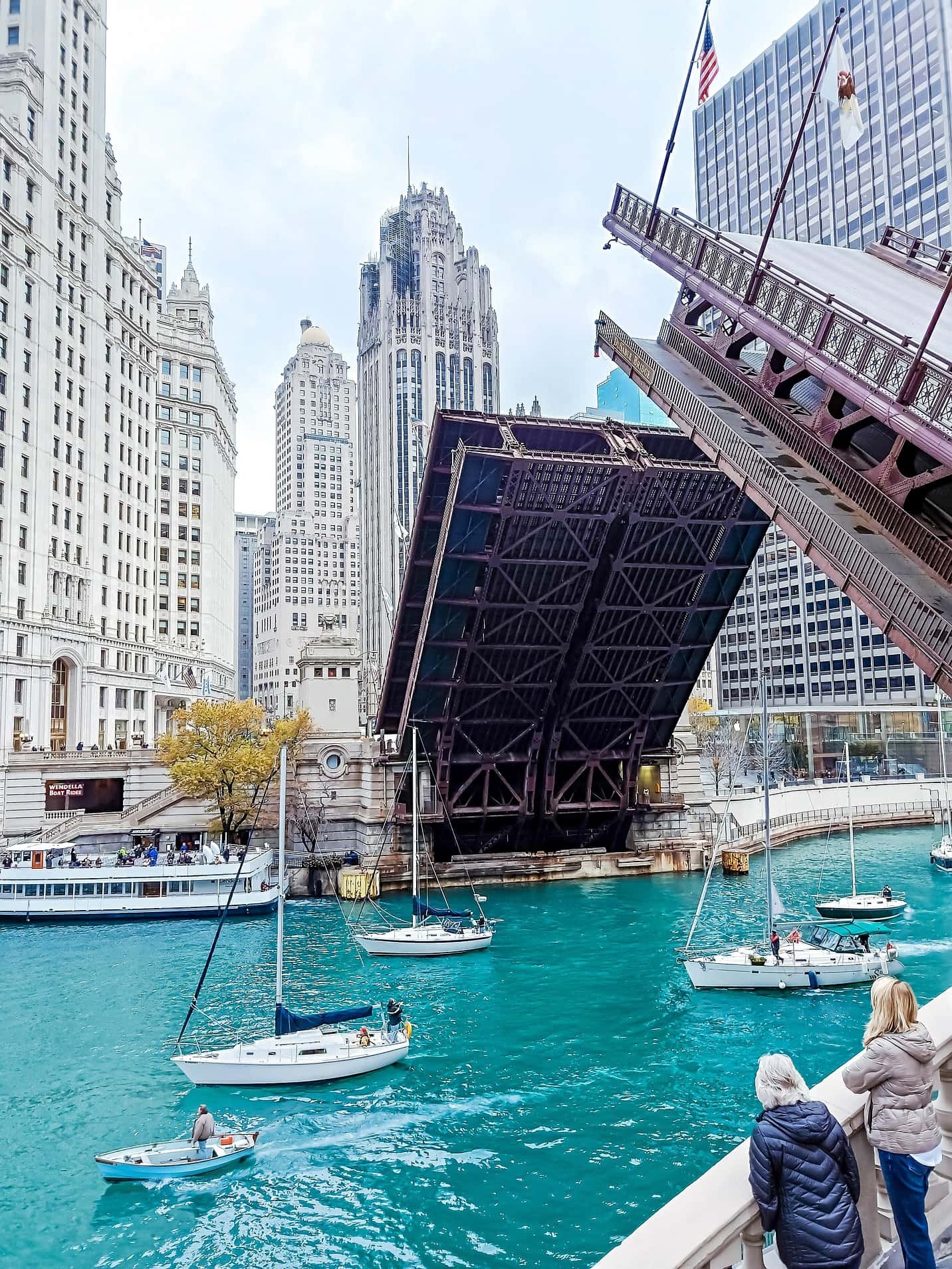 A raised bascule bridge over the Chicago river, sailboats moored below and towers rising behind.