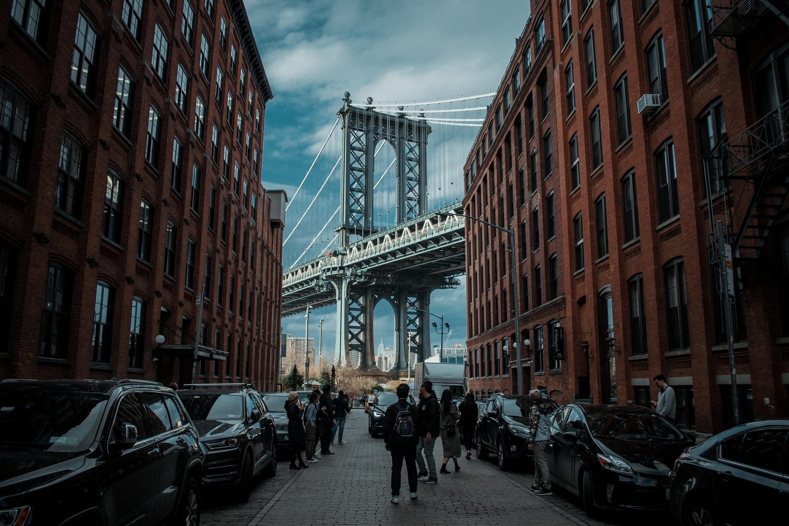 A steel suspension bridge framed between brick warehouses, with people walking the cobbled street below.