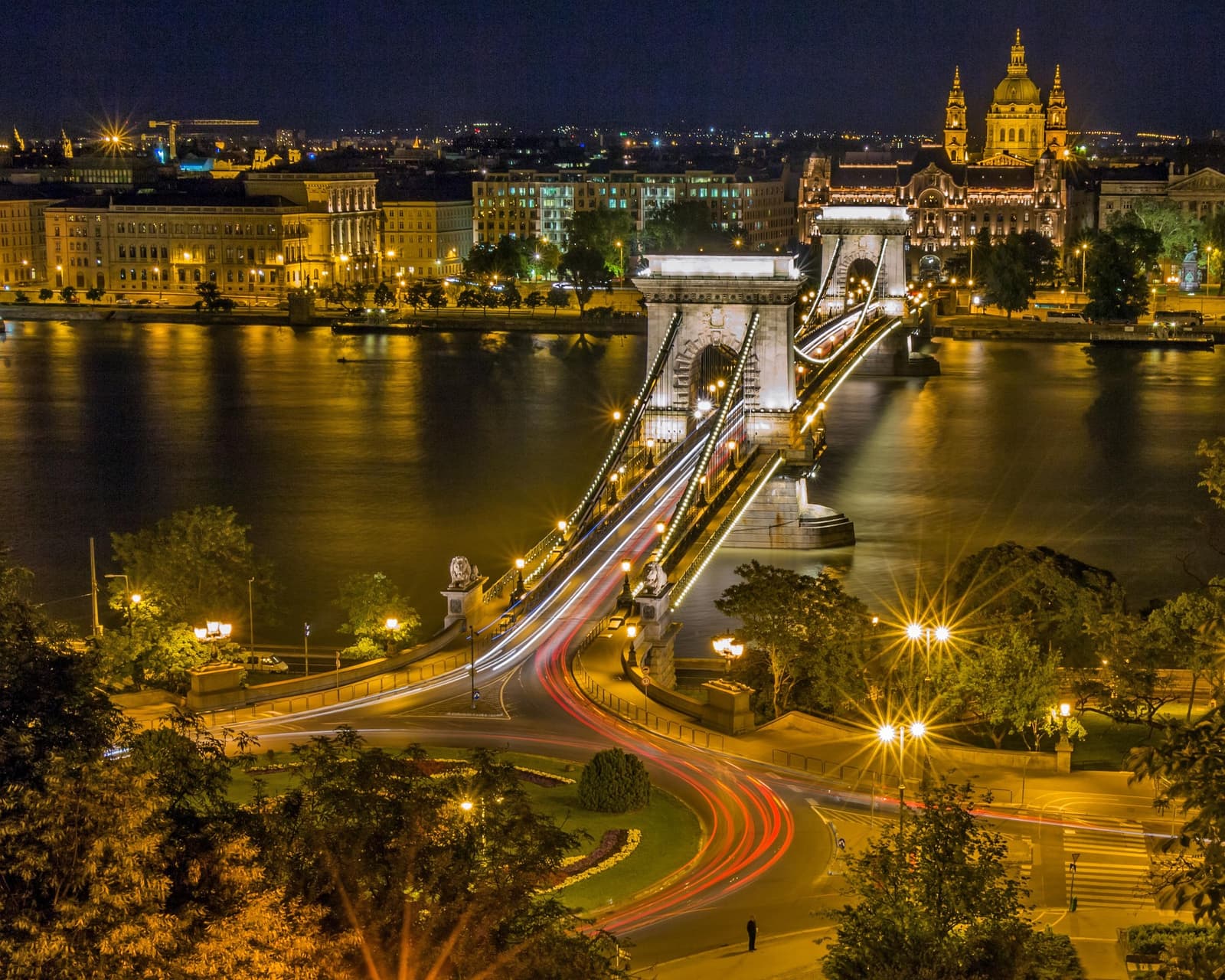 A lit chain bridge crossing a river at night, with a domed basilica on the skyline behind.